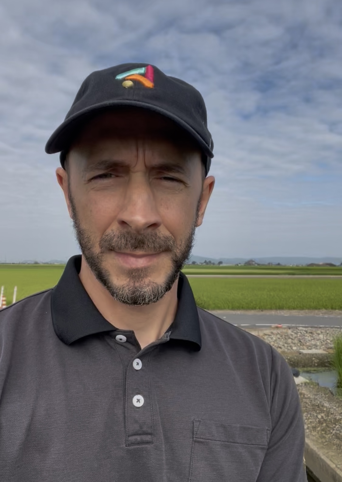 Me in a black cap and gray polo standing in front of green rice fields near my house in Japan.