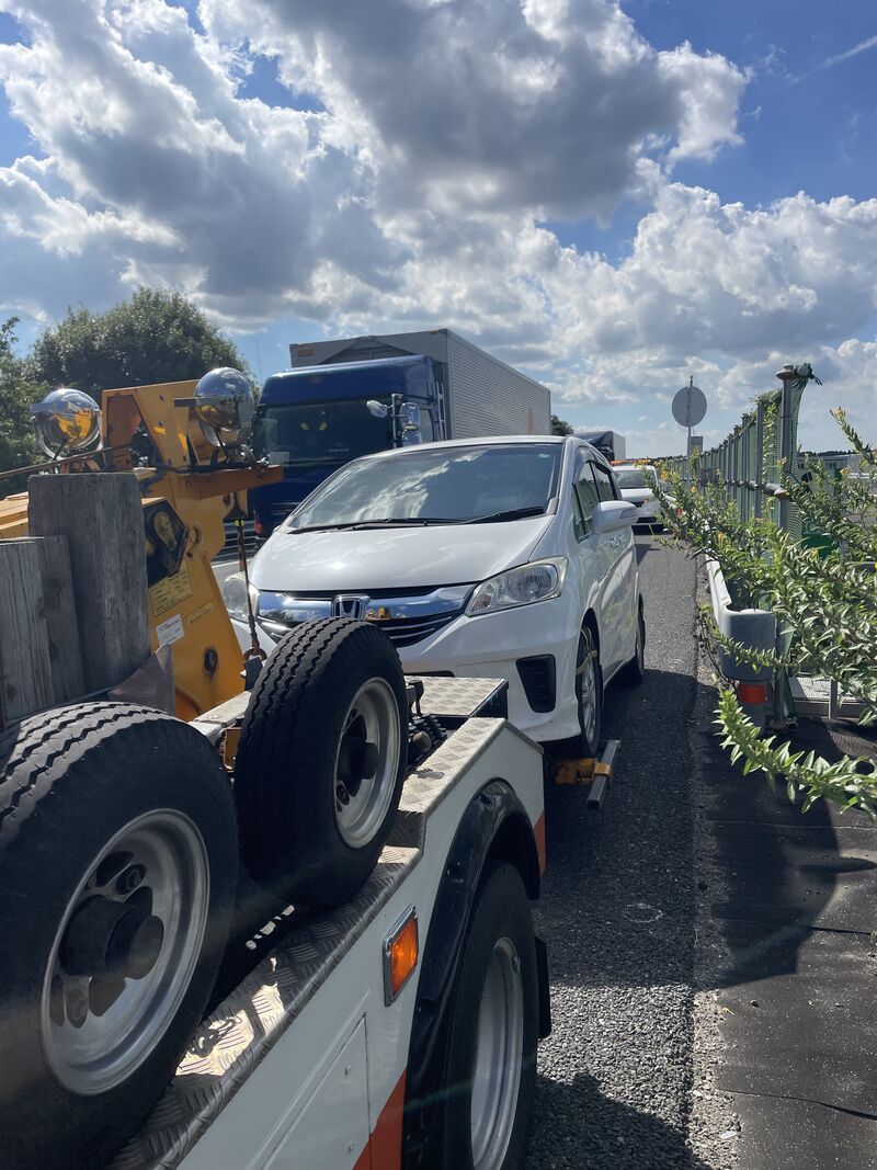 A white Honda minivan loaded onto the back of a yellow tow truck on the shoulder of a highway, with a blue truck passing and a construction fence to the right.