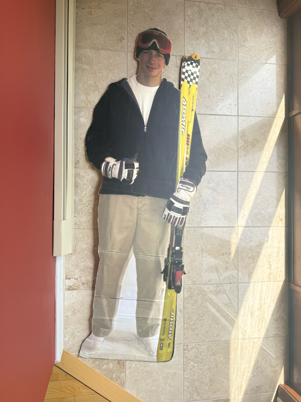 The life-sized cardboard cutout propped in a tiled hallway corner: a younger me in ski goggles, a dark half-zip over a white tee, khaki pants, and gloves, holding a pair of yellow Atomic skis.