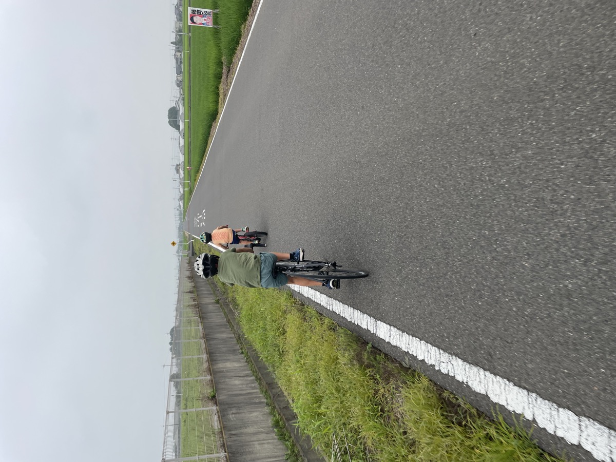 Two boys on bikes riding down a straight road between rice fields on an overcast day.