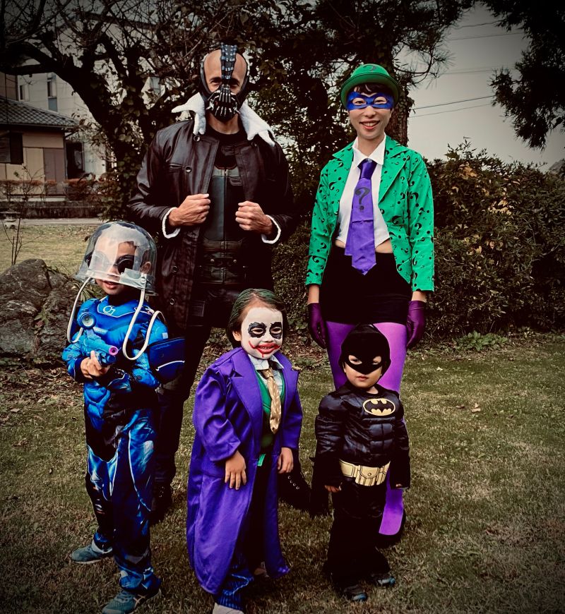 Our family in Batman costumes on the lawn: me as Bane in a leather jacket and mask, my wife as the Riddler in a green question-mark jacket, and the kids as a blue astronaut, the Joker, and Batman.