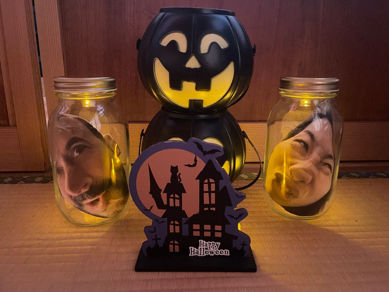 Halloween decorations on a tatami floor: two glowing mason jars with photos of a man's and a woman's faces pressed inside, a stacked jack-o'-lantern bucket, and a Happy Halloween haunted house sign.