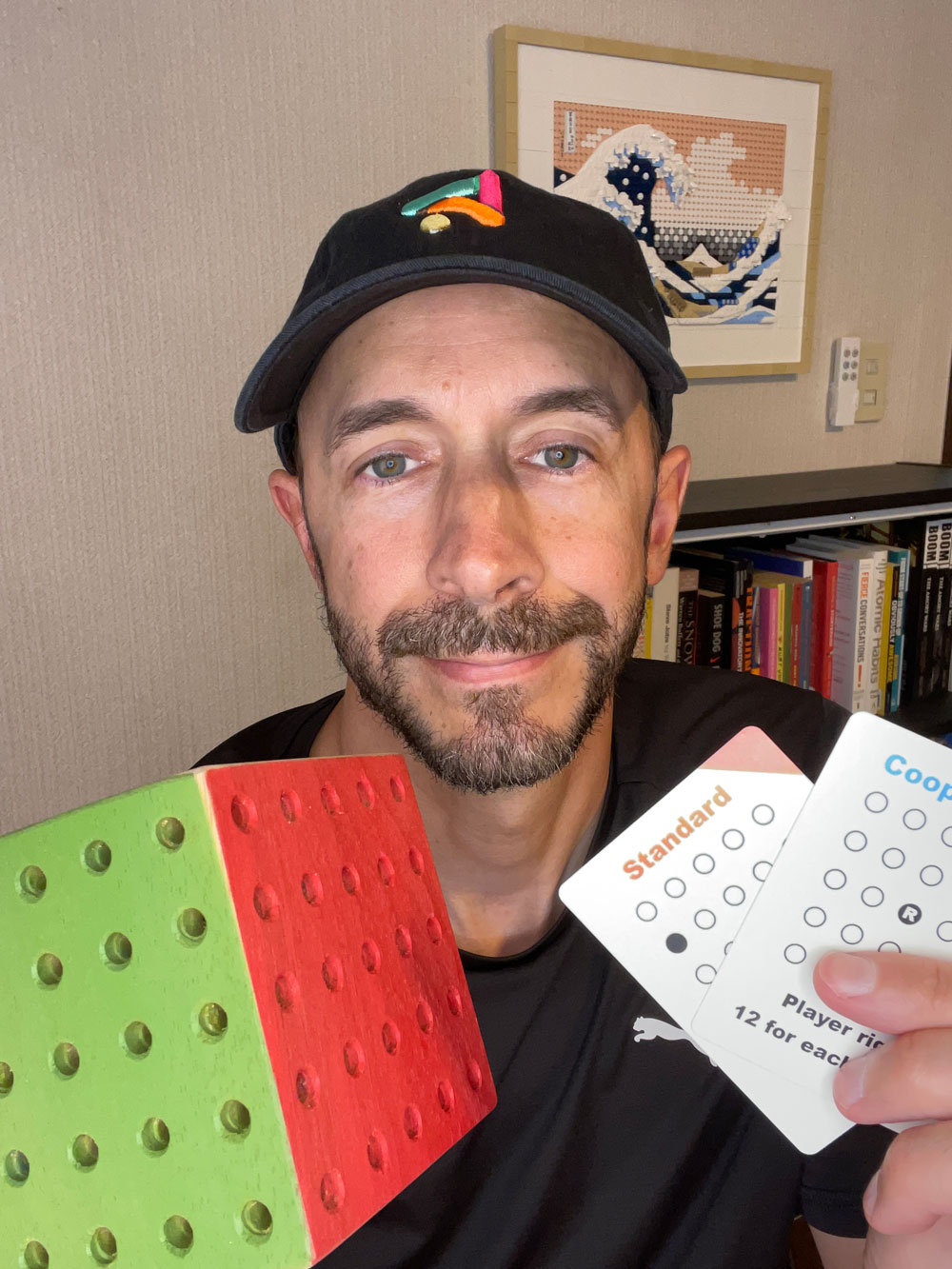 A selfie of me in a black cap and t-shirt holding up a hand-made wooden game board, painted green on one half and red on the other and covered in rows of pegs, next to two game cards labeled 'Standard' and 'Coop'. A framed Lego version of Hokusai's Great Wave and a bookshelf are behind me.