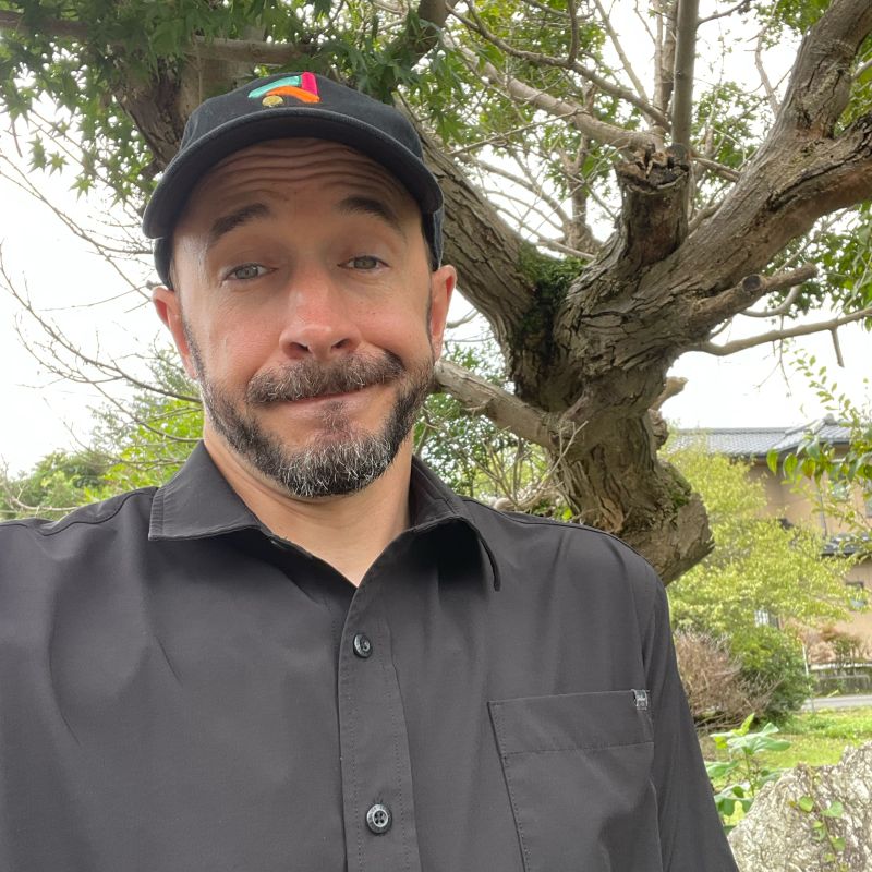 Kevin Dias in a black cap and black button-up shirt, shrugging with a wry smile in front of a tree.