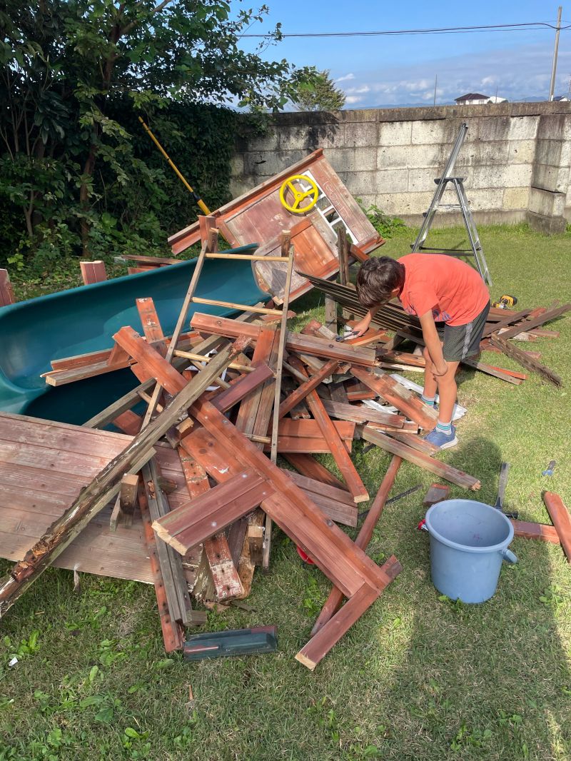 A pile of dismantled red playground lumber on the lawn, with a green slide, a yellow steering wheel, a stepladder, and my son in an orange shirt pulling boards from the heap.