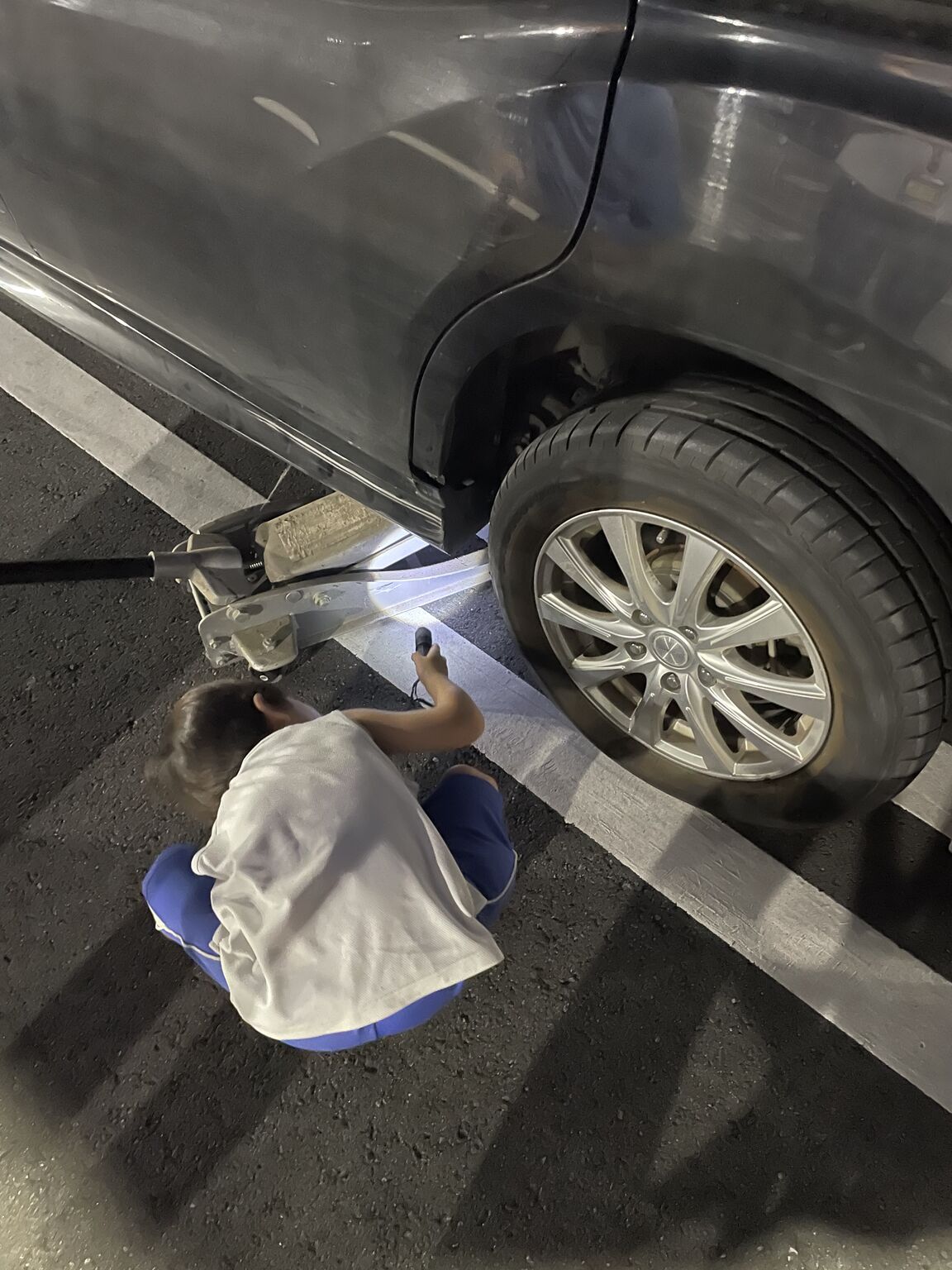 My four-year-old crouched next to the rear wheel of our car at night in a parking lot, shining a small flashlight at the jack under the frame.