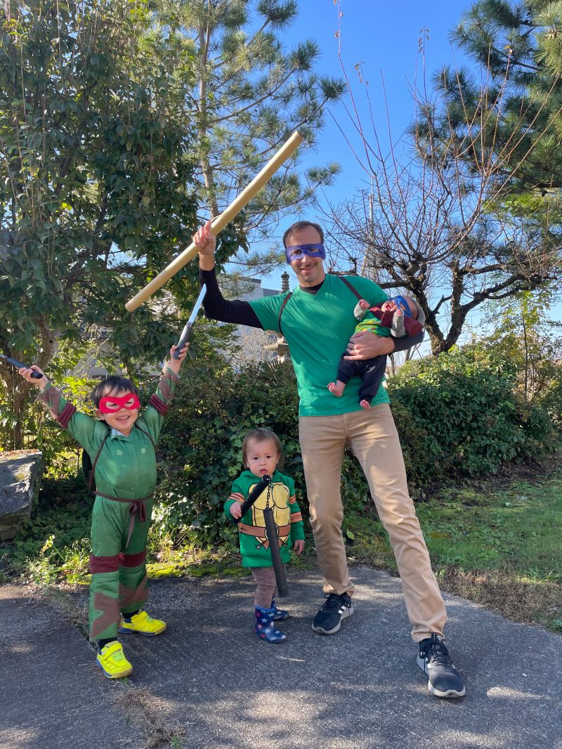Me as Donatello in a green shirt and purple mask, raising a bo staff and holding the baby in a turtle outfit, with the boys dressed as Raphael and a toddler turtle holding toy weapons.