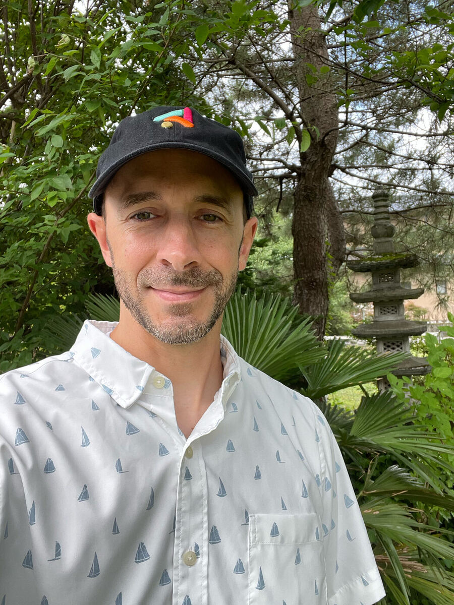 A selfie outdoors in a Japanese-style garden: me in a black cap with a colorful embroidered design and a white short-sleeve shirt patterned with sailboats, a stone lantern and greenery behind me.