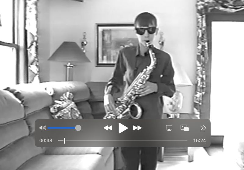 Black and white still from a digitized VHS tape of a 12-year-old boy in sunglasses playing a saxophone in a living room, with video player controls showing 00:38 of 15:24.