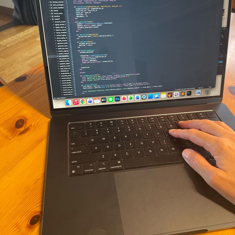 A laptop on a wooden table showing Ruby code in a text editor, with a hand resting on the keyboard.
