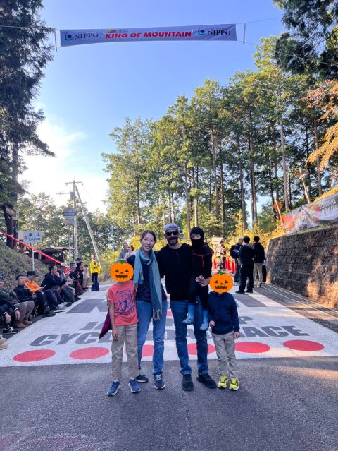 Kevin and his family standing on the painted road beneath the King of Mountain banner at the Japan Cup cycle road race, with spectators lining the roadside and cedar trees behind them.