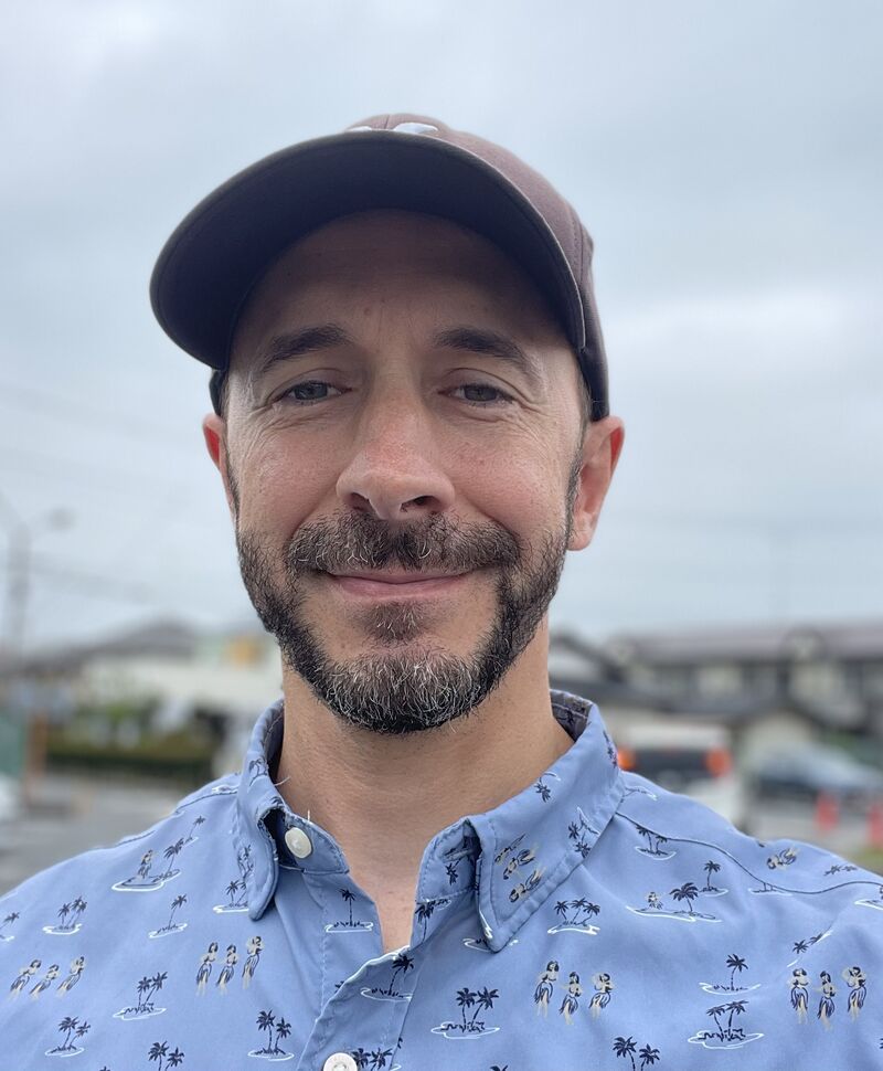 A selfie of me smiling in a brown baseball cap and a light blue shirt printed with palm trees and hula dancers, with houses and a cloudy sky in the background.