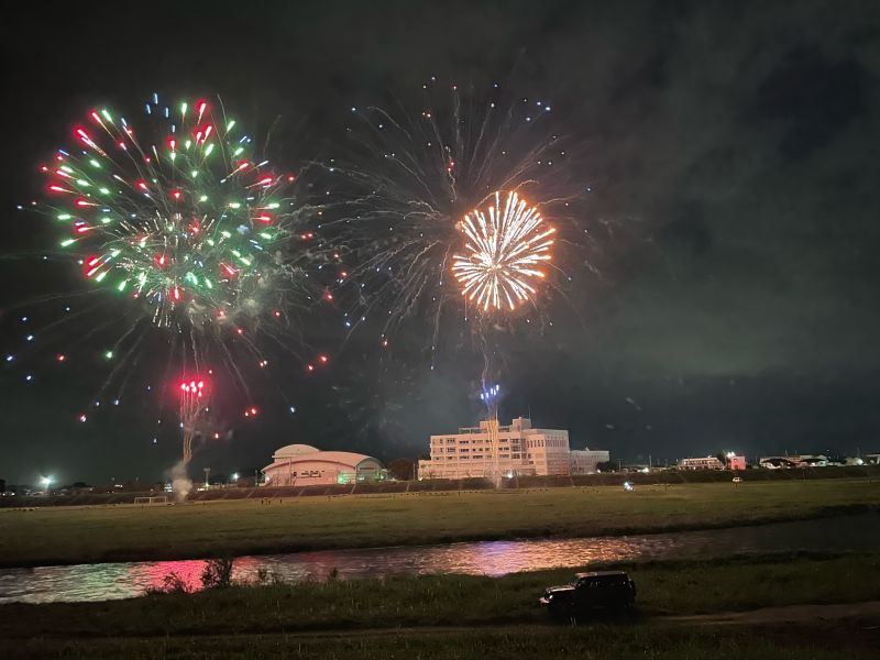 Red, green, and gold fireworks bursting over the river in Oyama, with their reflection in the water.