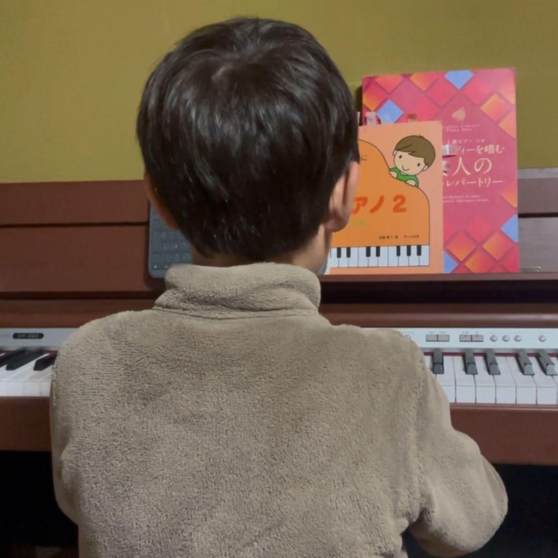 My son, seen from behind in a tan fleece, sitting at a digital piano with Japanese piano method books propped on the music stand.