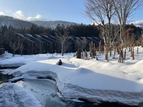 Snow-covered grounds of a hotel in Nagano, with dark wooden lodge buildings, a steaming hot spring stream winding through deep snow, bare trees, and mountains under a blue sky.