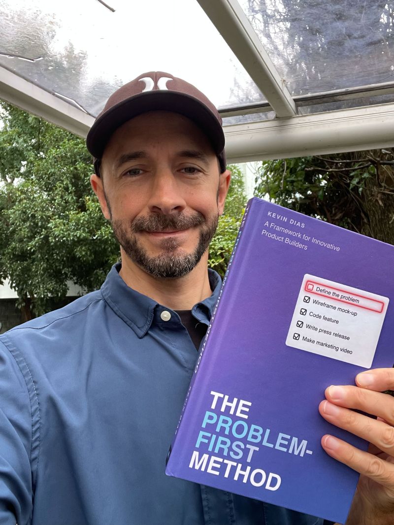 Kevin Scott Dias standing under a rain-streaked glass roof in a brown cap and blue shirt, holding up a purple hardcover copy of The Problem-First Method.