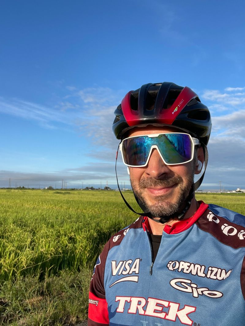 A selfie in a cycling helmet, sunglasses, and a Trek jersey in front of a bright green rice field under a blue sky.