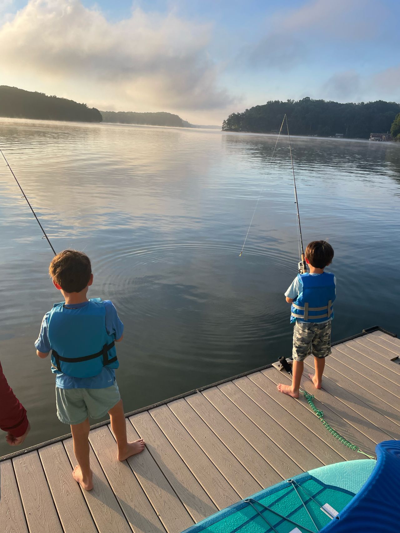 Two boys in blue life jackets fishing off a dock on a calm lake, with morning mist rising off the water.