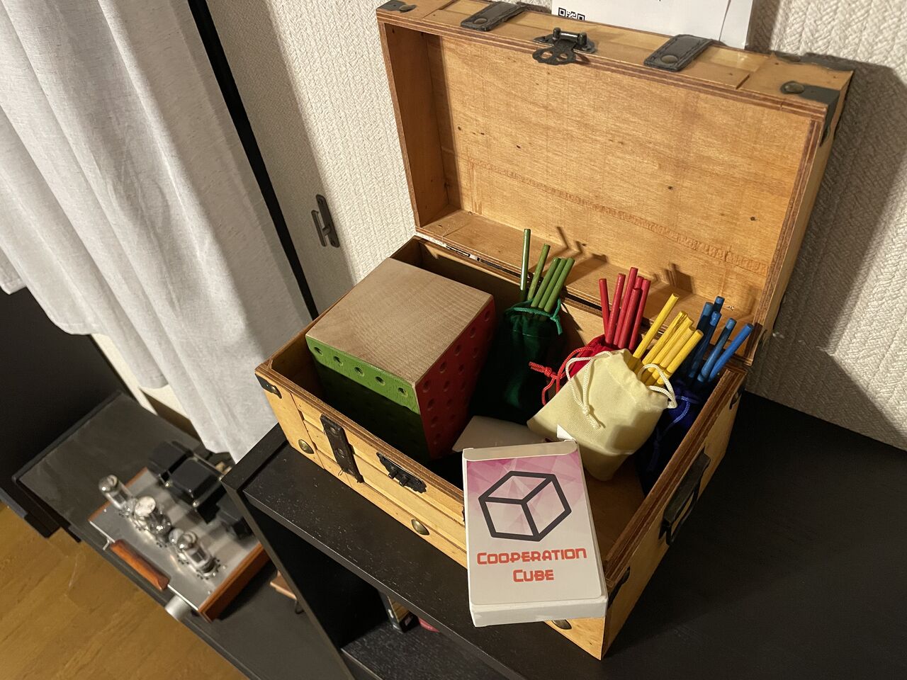 A handmade Cooperation Cube set inside an open wooden chest: a wooden cube with colored pegboard faces, cloth bags of colored pegs, and a deck of Cooperation Cube cards.
