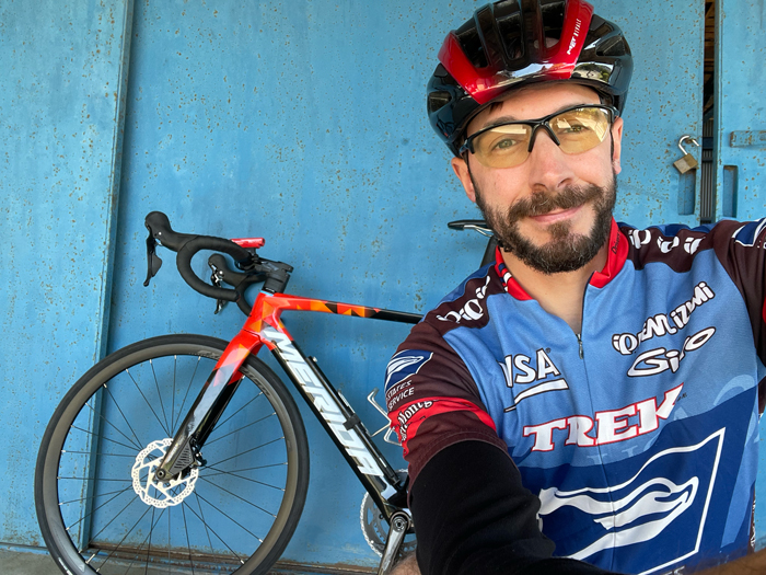 A cycling selfie of me in a red and black helmet and yellow-tinted glasses, wearing a Trek team jersey, beside my orange and black Merida road bike against a blue garage door.