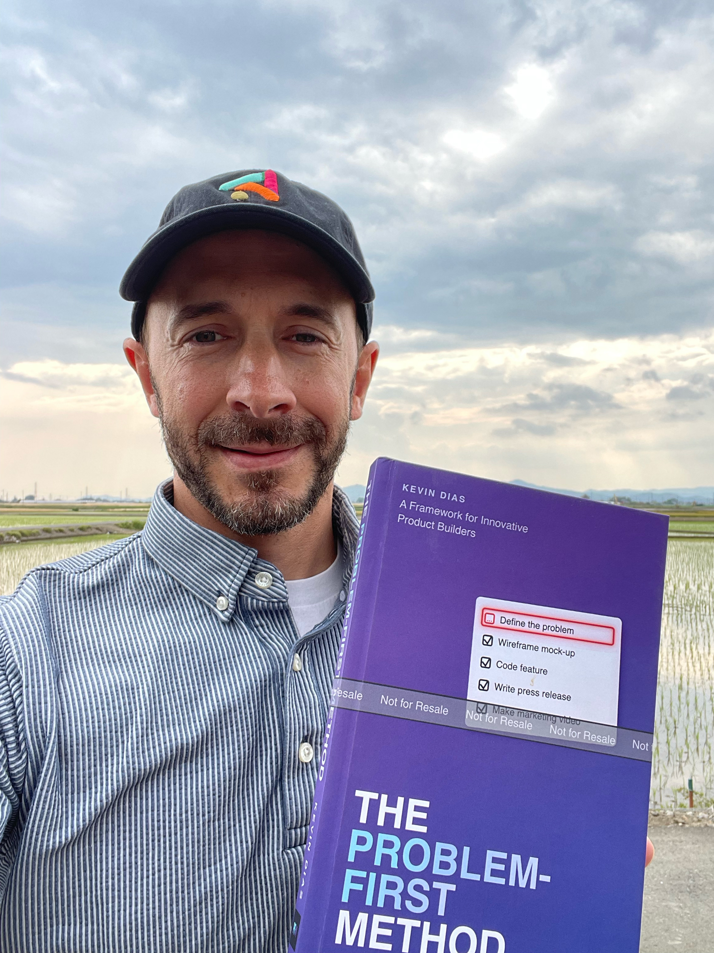 Kevin Dias in a cap and striped shirt holding a proof copy of The Problem-First Method in front of a rice paddy under a cloudy sky.