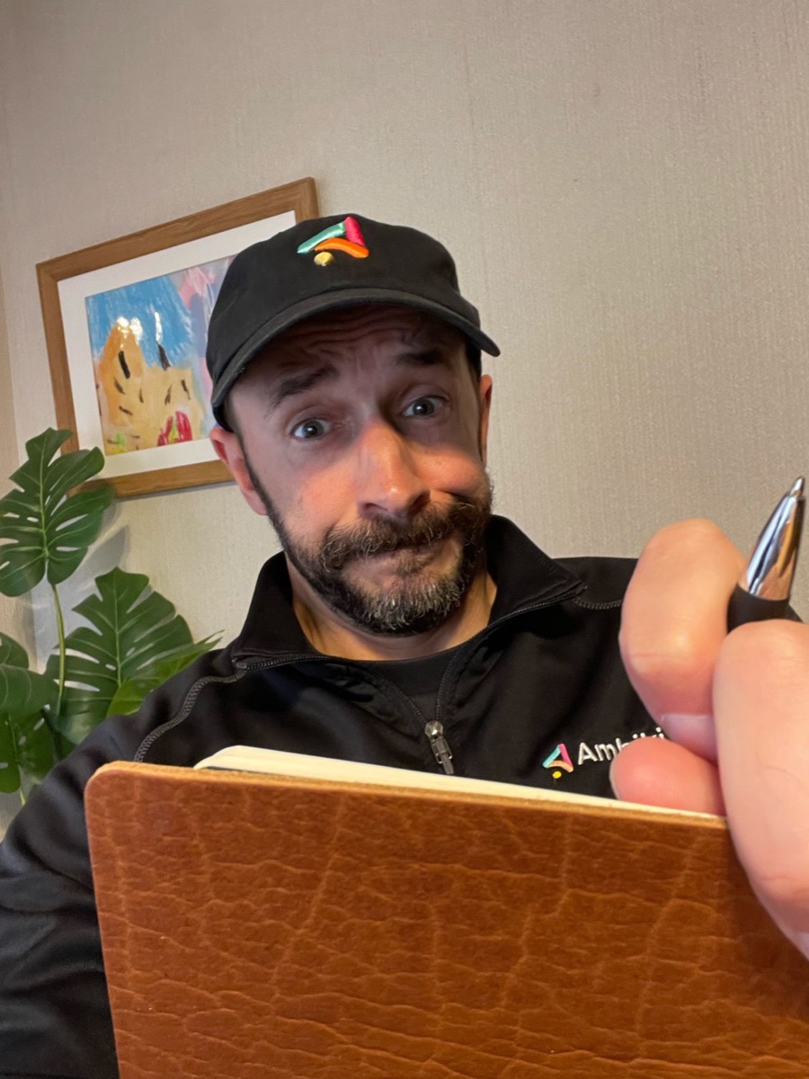 Kevin in a black Ambiki cap and jacket, eyebrows raised and lips pressed together, holding a pen over a brown leather notebook, with a child's framed painting and a leafy plant behind him.